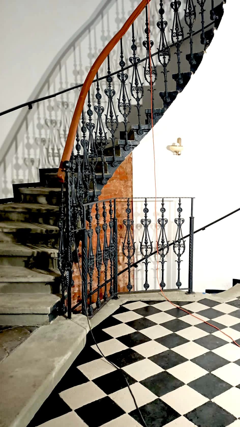 Klatka schodowa z metalową, zdobioną balustradą w stylu retro. Balustrada pomalowana na czarno, z drewnianą poręczą. Podłoga w czarno-białą szachownicę.