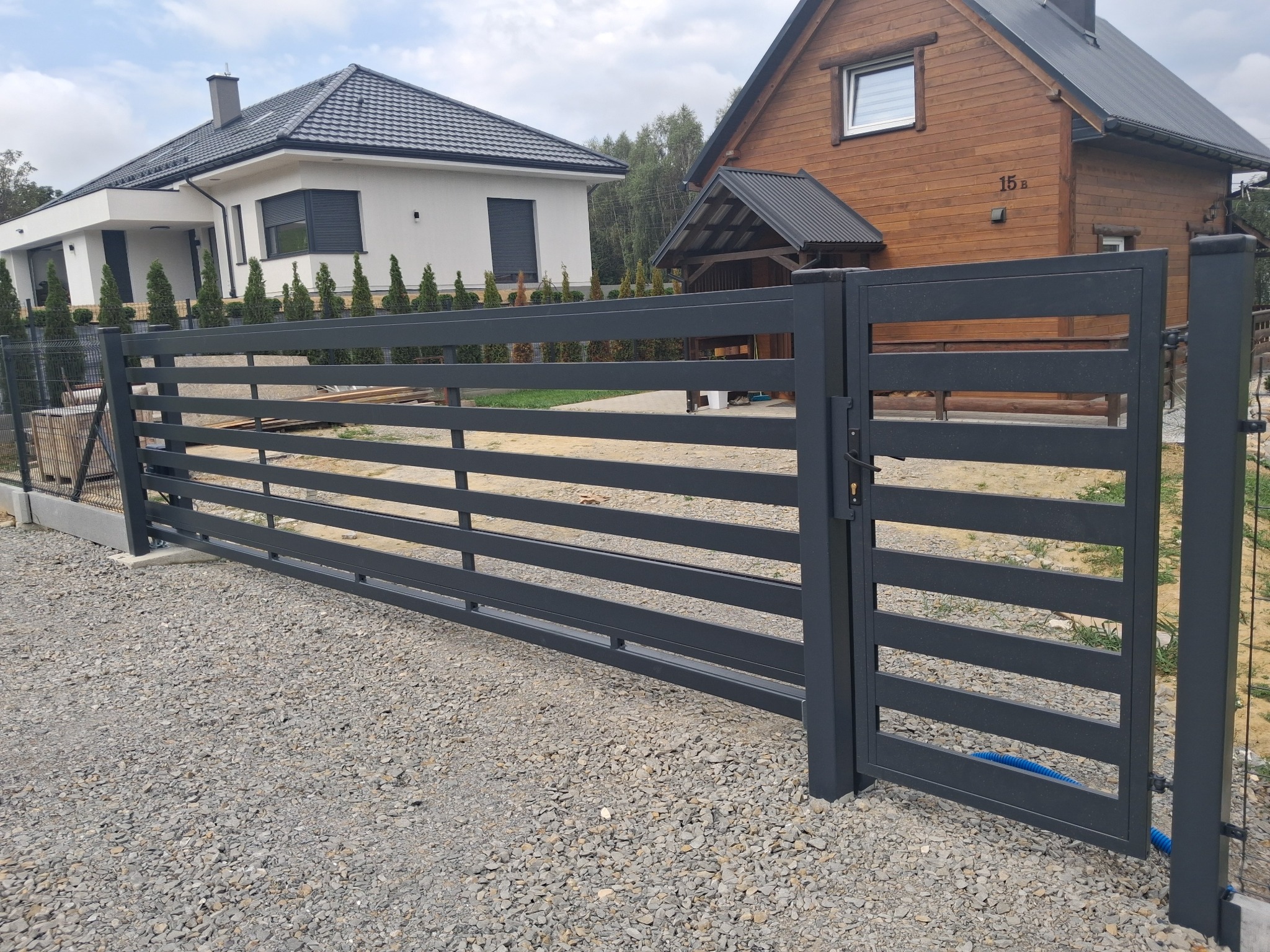 Nowoczesna, metalowa brama wjazdowa w kolorze grafitowym. Horyzontalne, płaskie profile. Domy w tle. Brama zamontowana na posesji z podłożem z kamyków.