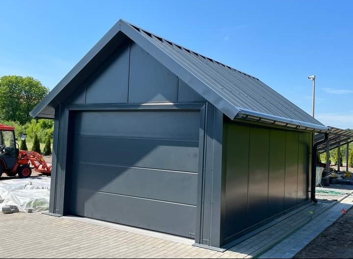 Nowoczesny, grafitowy garaż blaszany z bramą segmentową, na kostce brukowej, z traktorem w tle i widocznym systemem rynnowym.
