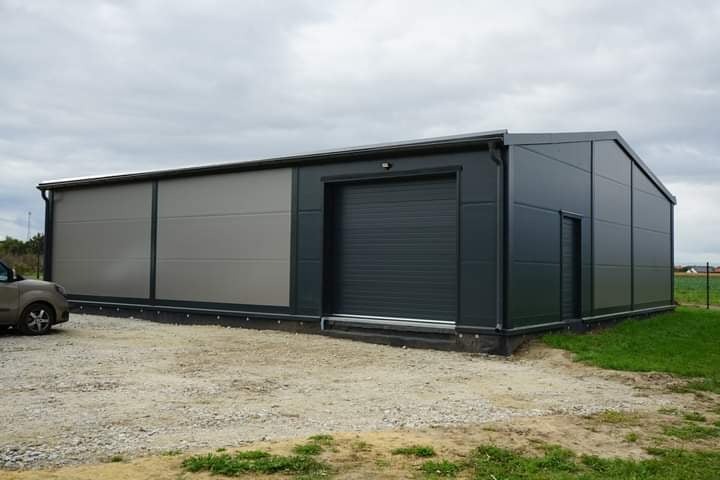 Nowoczesna hala z płyt warstwowych w odcieniach szarości, z bramą rolowaną i drzwiami wejściowymi, na żwirowym podłożu, pod pochmurnym niebem.