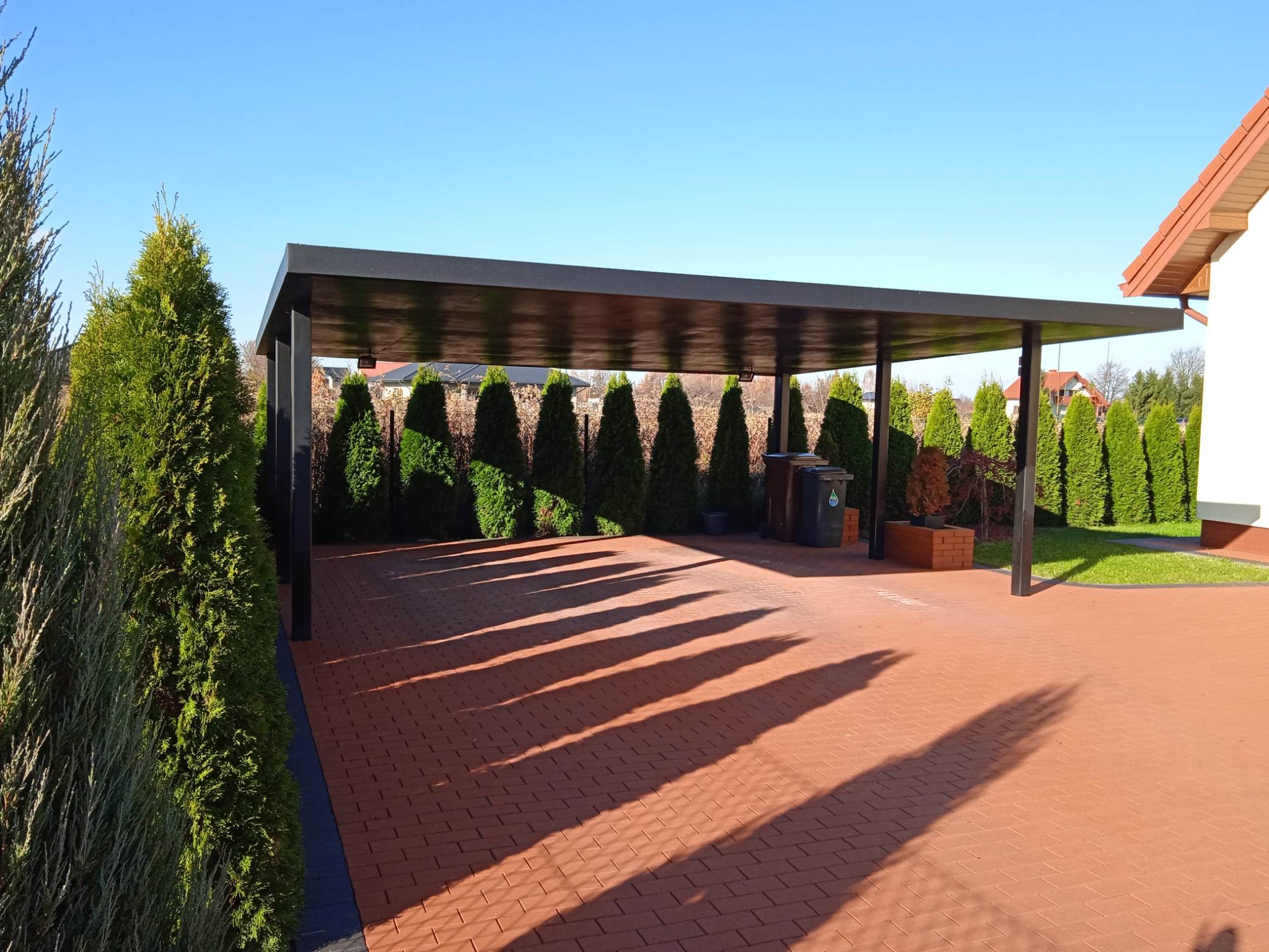 Nowoczesna, czarna pergola metalowa na posesji z kostką brukową i tujami, rzucająca długie cienie w słoneczny dzień. Minimalistyczny design.