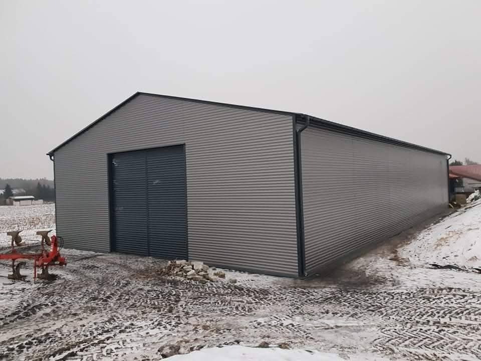 Szara hala z blachy falistej z ciemnymi drzwiami przesuwnymi, widoczna na tle zaśnieżonego pola i pochmurnego nieba. Na pierwszym planie resztki śniegu i ślady opon.