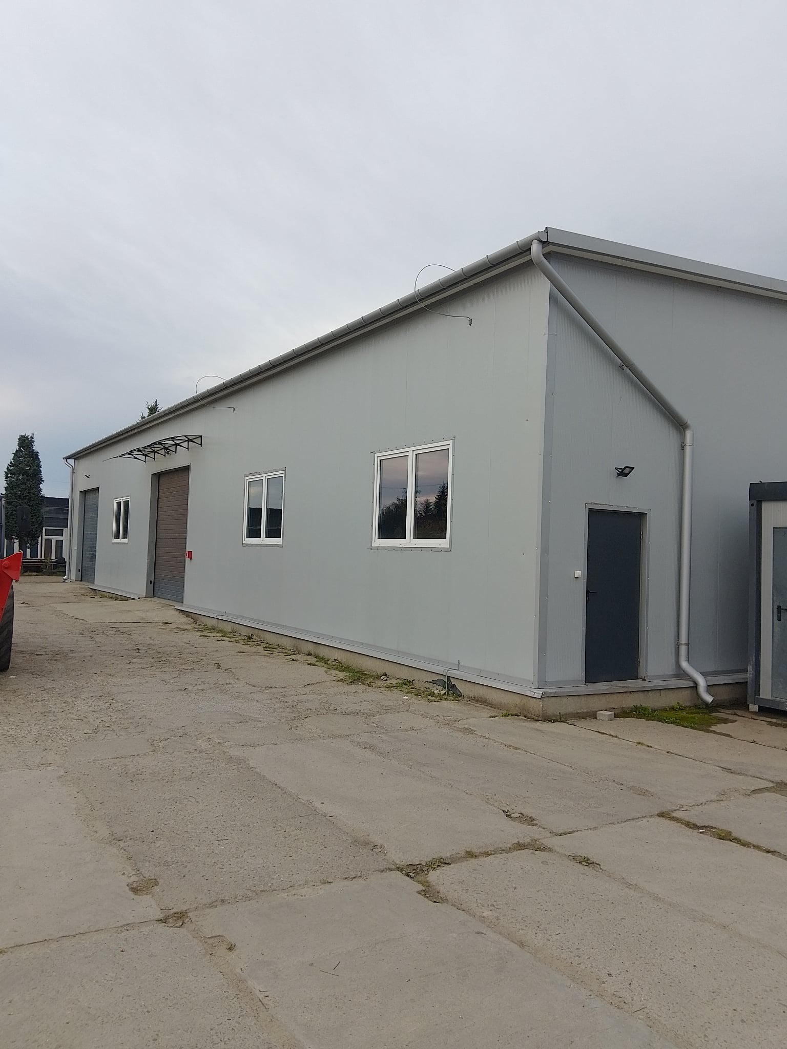 Szara hala z płyt warstwowych z oknami i drzwiami, widok z zewnątrz, betonowy plac przed budynkiem, pochmurne niebo.