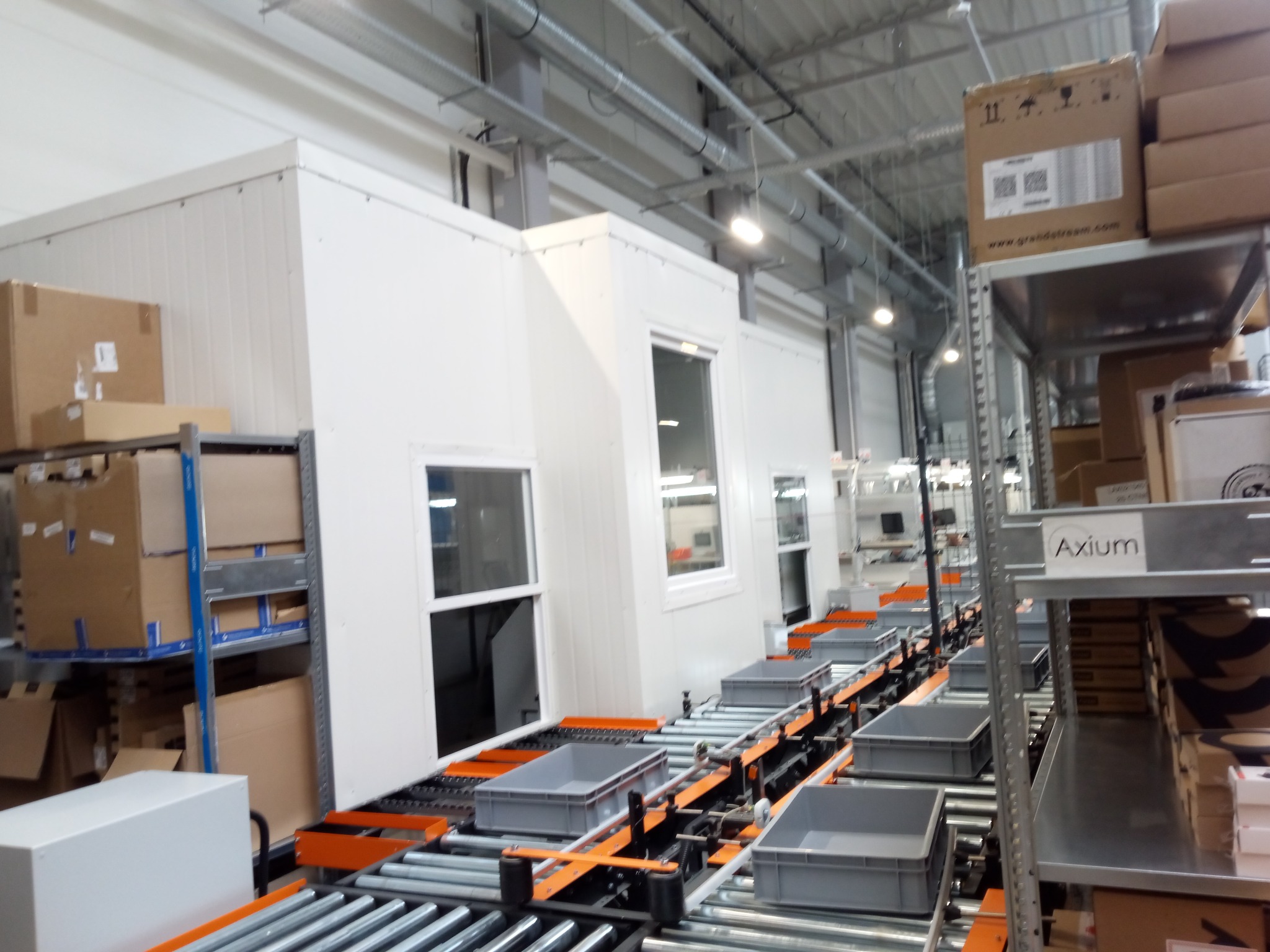Wnętrze hali produkcyjnej z linią transportową i modułowymi pomieszczeniami. Widoczne regały z kartonami i elementy konstrukcji dachu. Oświetlenie przemysłowe.