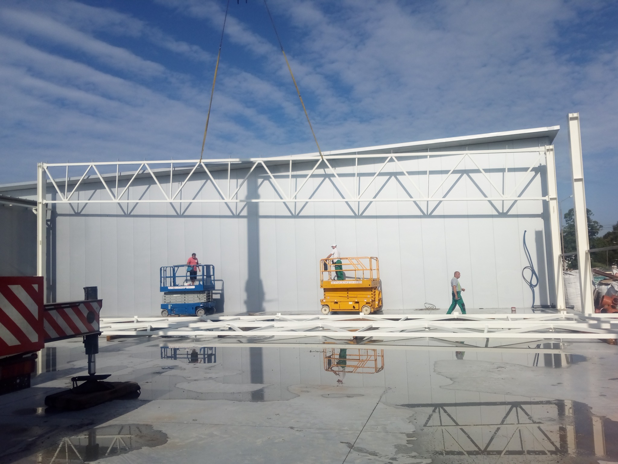 Montaż hali: pracownicy na podnośnikach instalują białe płyty warstwowe. Na mokrej posadzce leżą elementy konstrukcji, odbijające się w kałużach. Widoczne dźwigi.