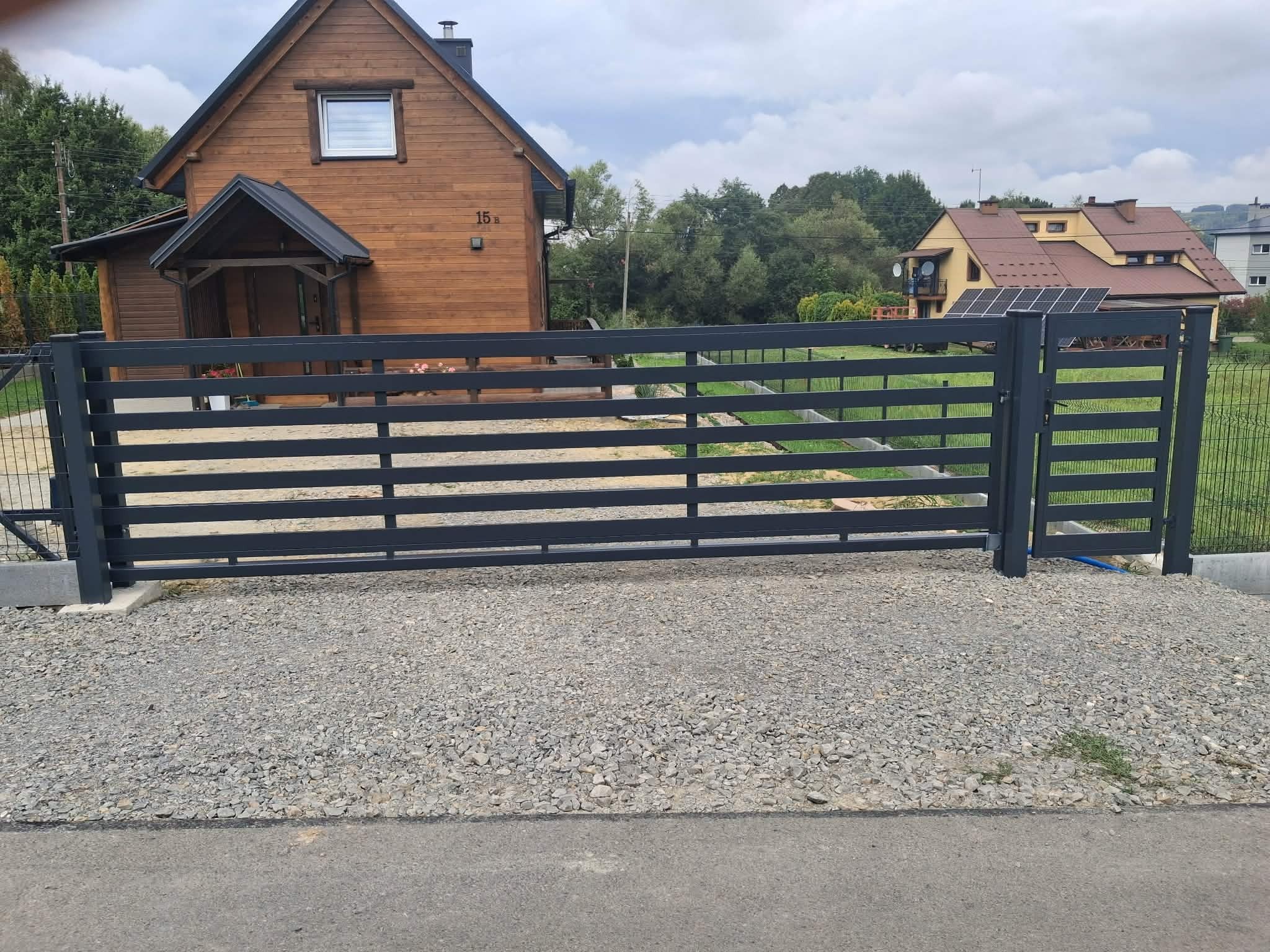 Nowoczesna, pozioma brama wjazdowa w kolorze antracytowym, na tle drewnianego domu z numerem 15b i zieleni. Brama zamontowana na żwirowym podłożu.