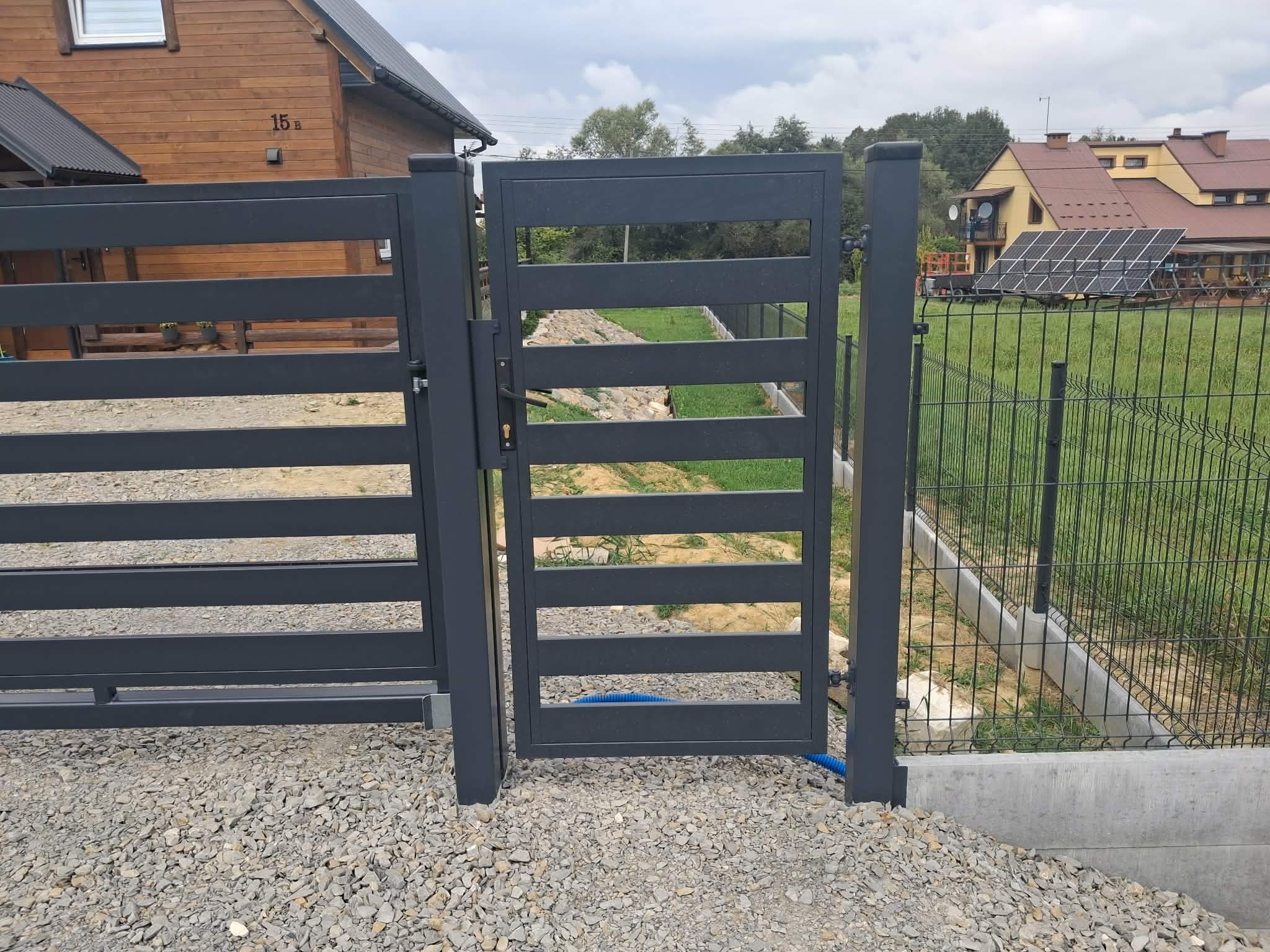 Nowoczesna, grafitowa brama wjazdowa z poziomymi szczeblami, otwierana na posesję z domem i panelami słonecznymi w tle. Widoczny fragment niebieskiego węża.