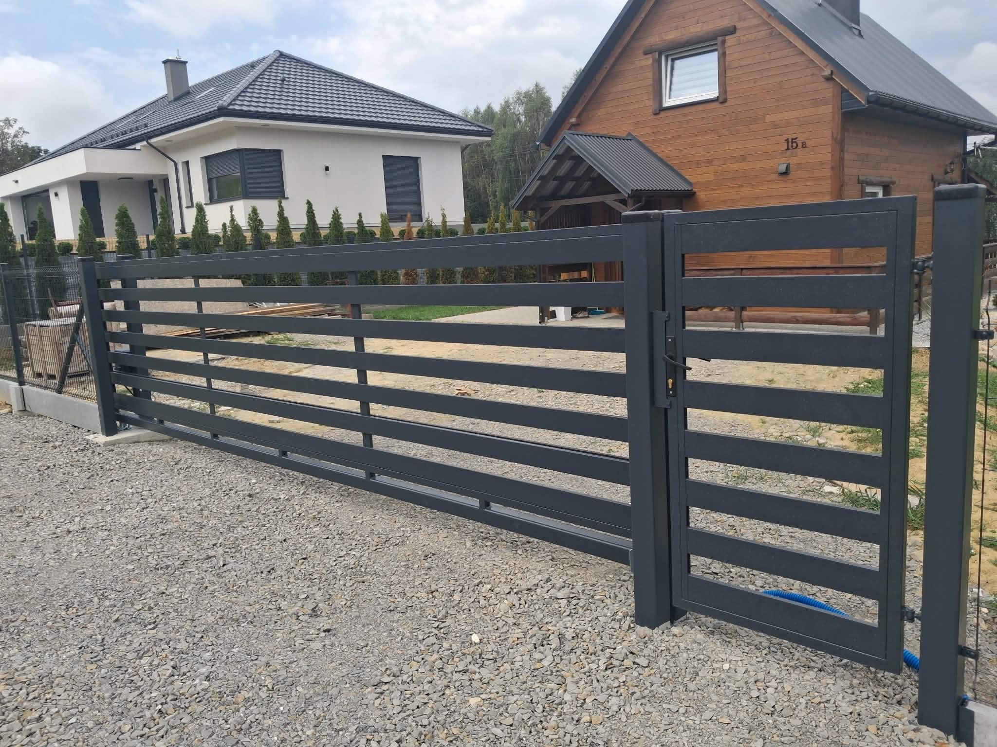 Nowoczesna, pozioma brama wjazdowa w kolorze grafitowym, z widocznymi dwoma domami w tle, jeden w stylu nowoczesnym, drugi drewniany. Brama na żwirowym podłożu.