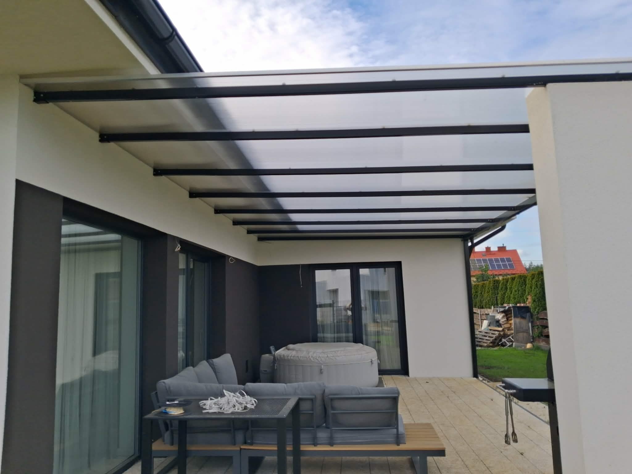 Nowoczesna pergola z czarną konstrukcją i przezroczystym zadaszeniem, osłaniająca taras z meblami ogrodowymi i jacuzzi. Widok na dom z panelami słonecznymi.