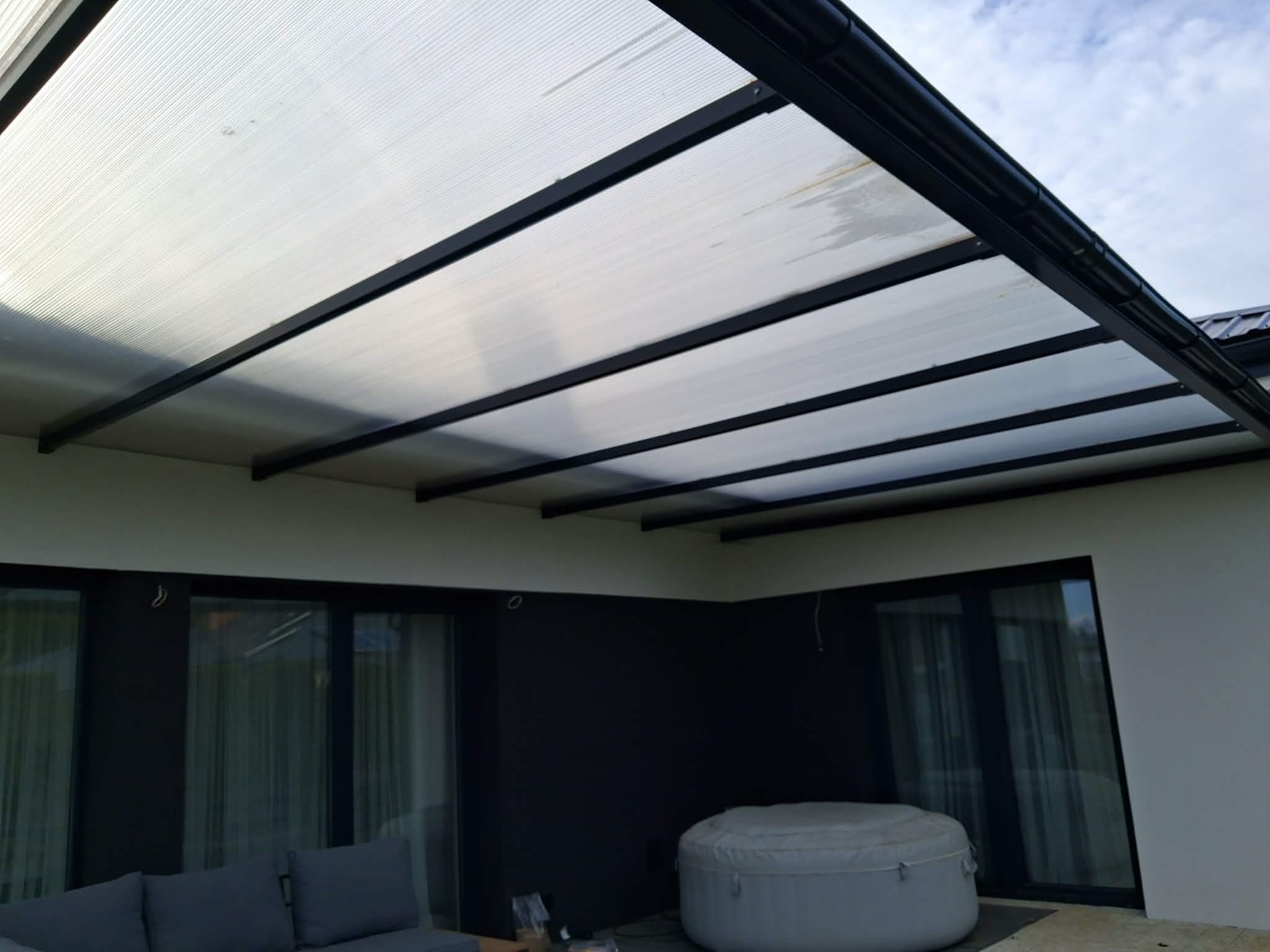 Nowoczesna pergola z czarną metalową konstrukcją i przezroczystym zadaszeniem, chroniąca taras z meblami ogrodowymi i jacuzzi. Widok z dołu, jasne niebo w tle.