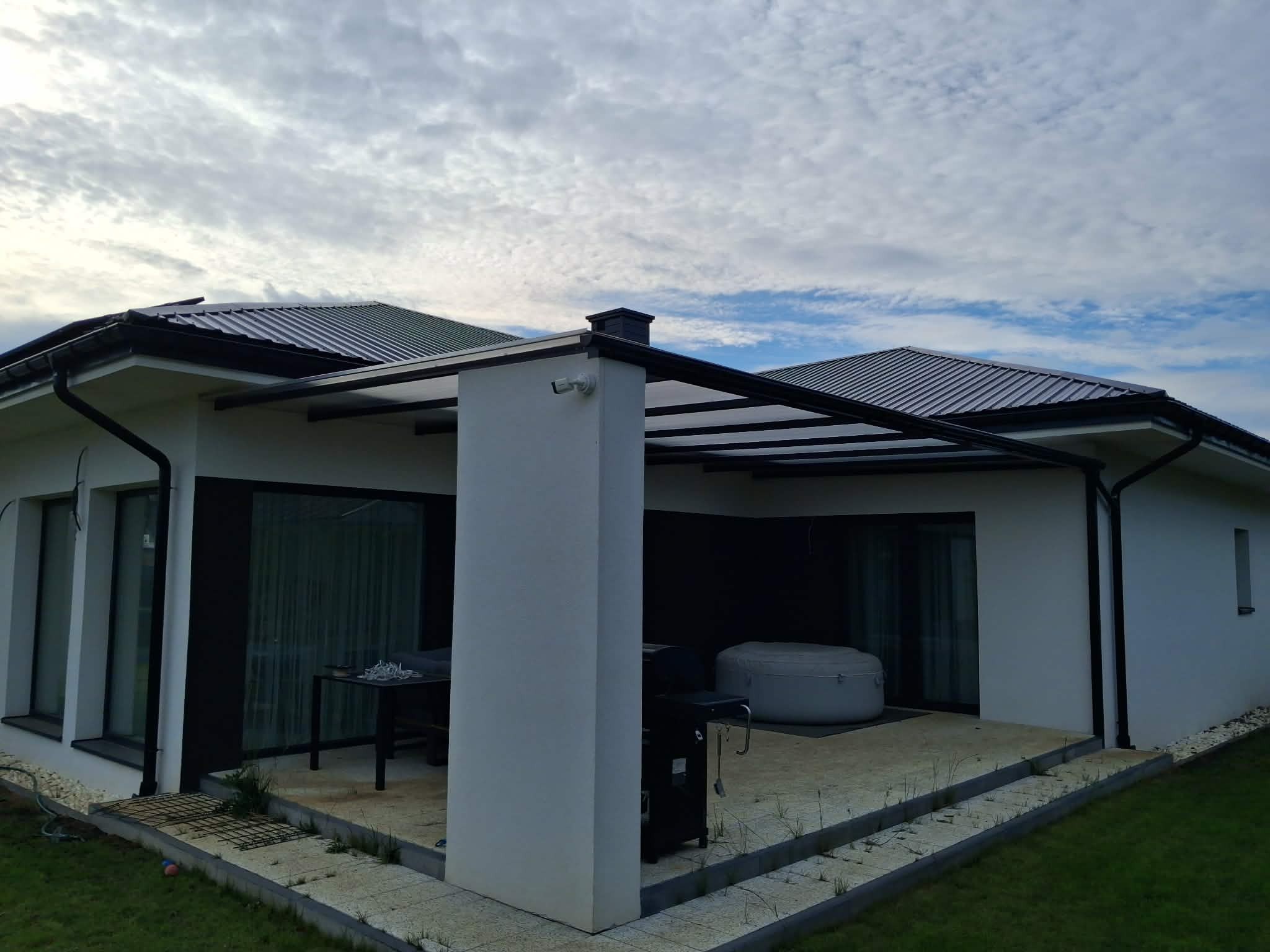 Nowoczesna pergola metalowa z zadaszeniem przy białym domu jednorodzinnym. Widoczne elementy konstrukcyjne i zadaszenie. W tle pochmurne niebo.