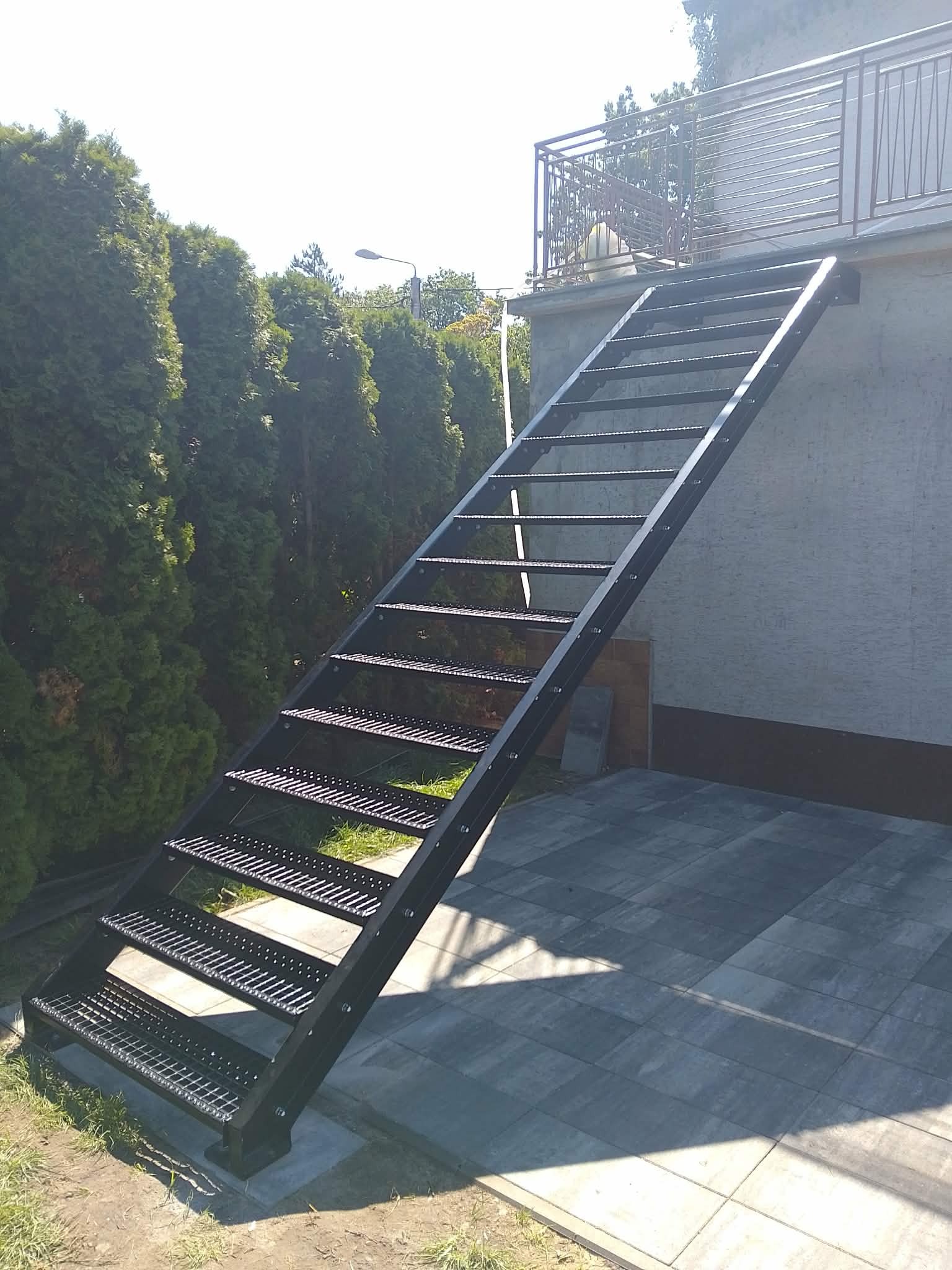 Zewnętrzne, metalowe schody kratowe prowadzące na balkon z balustradą, na tle zielonych drzew i szarej elewacji budynku. Widok z dołu, w słoneczny dzień.