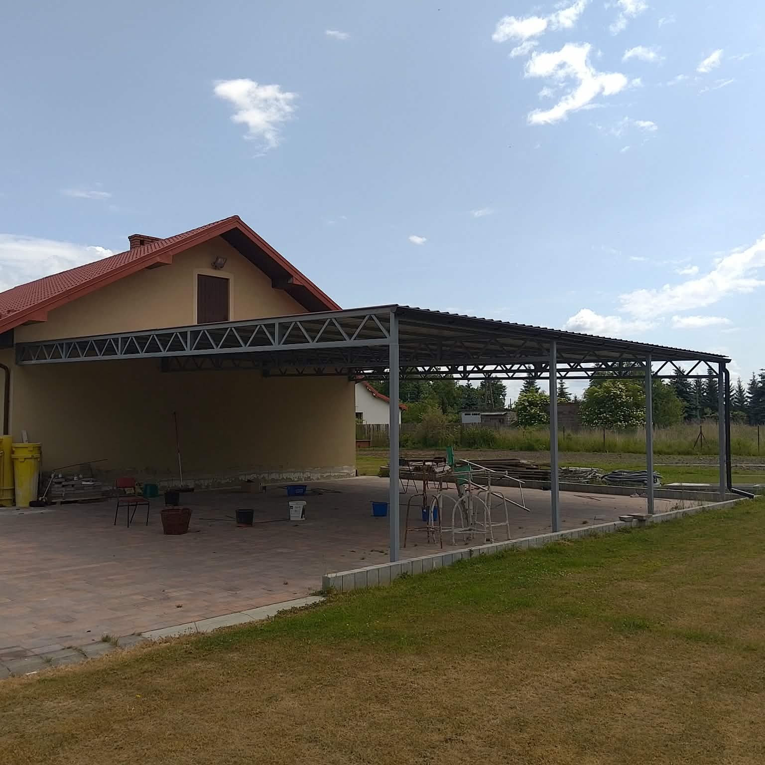 Zadaszenie tarasu z metalową konstrukcją przy domu jednorodzinnym. Widoczne elementy konstrukcyjne i zarys dachu. Tło: błękitne niebo z chmurami.