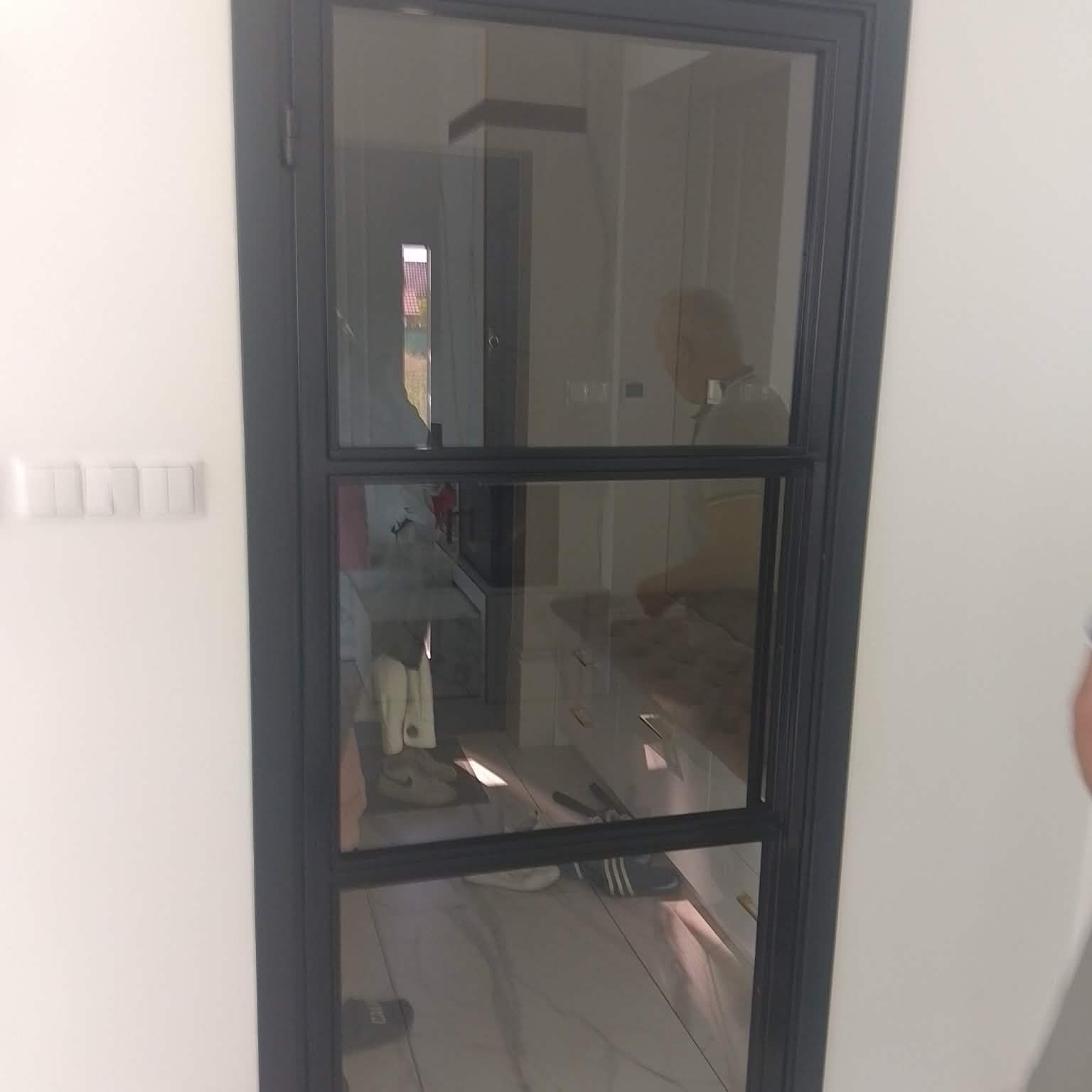 Nowoczesne, czarne drzwi metalowe z szybą, podzielone na trzy sekcje, w jasnym wnętrzu. Widoczne odbicie osoby i wyposażenia.