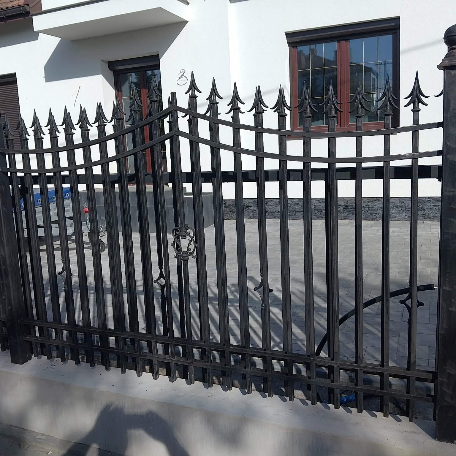 Czarna, metalowa brama wjazdowa z dekoracyjnymi grotami na tle białego domu z brązowymi oknami. Widoczne cieniowanie i kostka brukowa.
