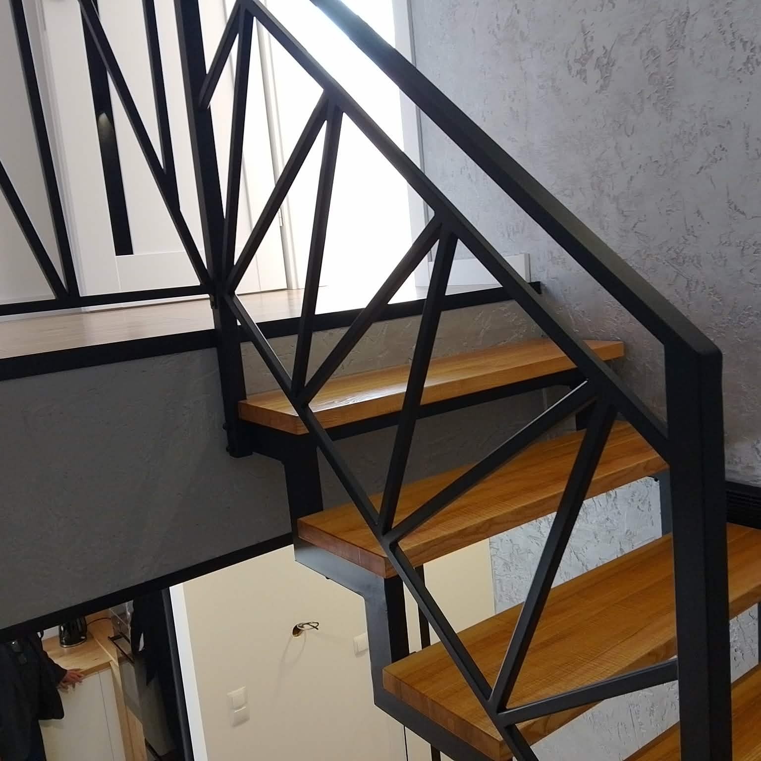 Nowoczesne schody metalowe z drewnianymi stopniami, czarna geometryczna balustrada, widok z góry, jasne wnętrze, minimalistyczny design.