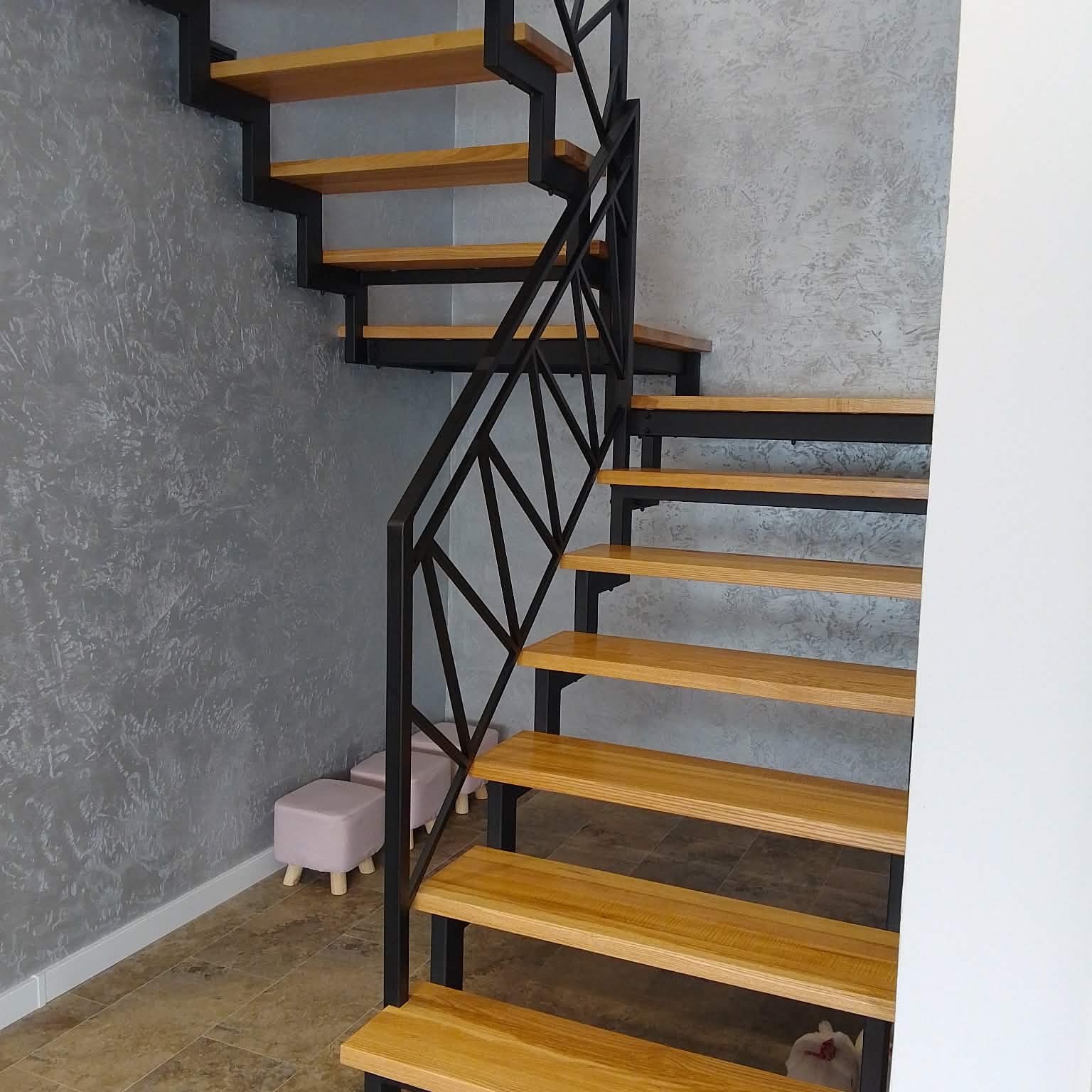 Nowoczesne schody metalowe z drewnianymi stopniami i geometryczną balustradą na tle szarej ściany z fakturą. Widoczne dwa pufy i fragment podłogi.