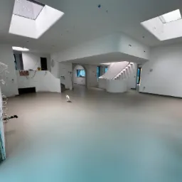 Jasnoszara, gładka posadzka w przestronnym, jasnym wnętrzu z oknami dachowymi. Widoczne schody, instalacje elektryczne i przygotowania do wykończenia.