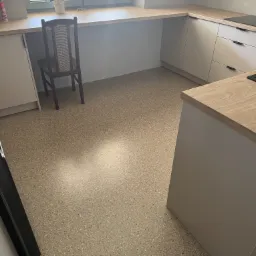 Jasna kuchnia z posadzką o wzorze lastryko. Krzesło przy oknie z kwiatami. Minimalistyczne szafki i blat imitujący drewno. Kontrastowe, czarne detale.