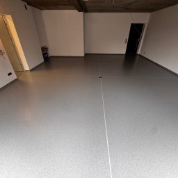 Tobiasz Krzak - Szara, gładka posadzka żywiczna w surowym wnętrzu z betonowym sufitem. Widoczne białe ściany i czarne drzwi. Minimalistyczny design, surowy stan wykończenia.