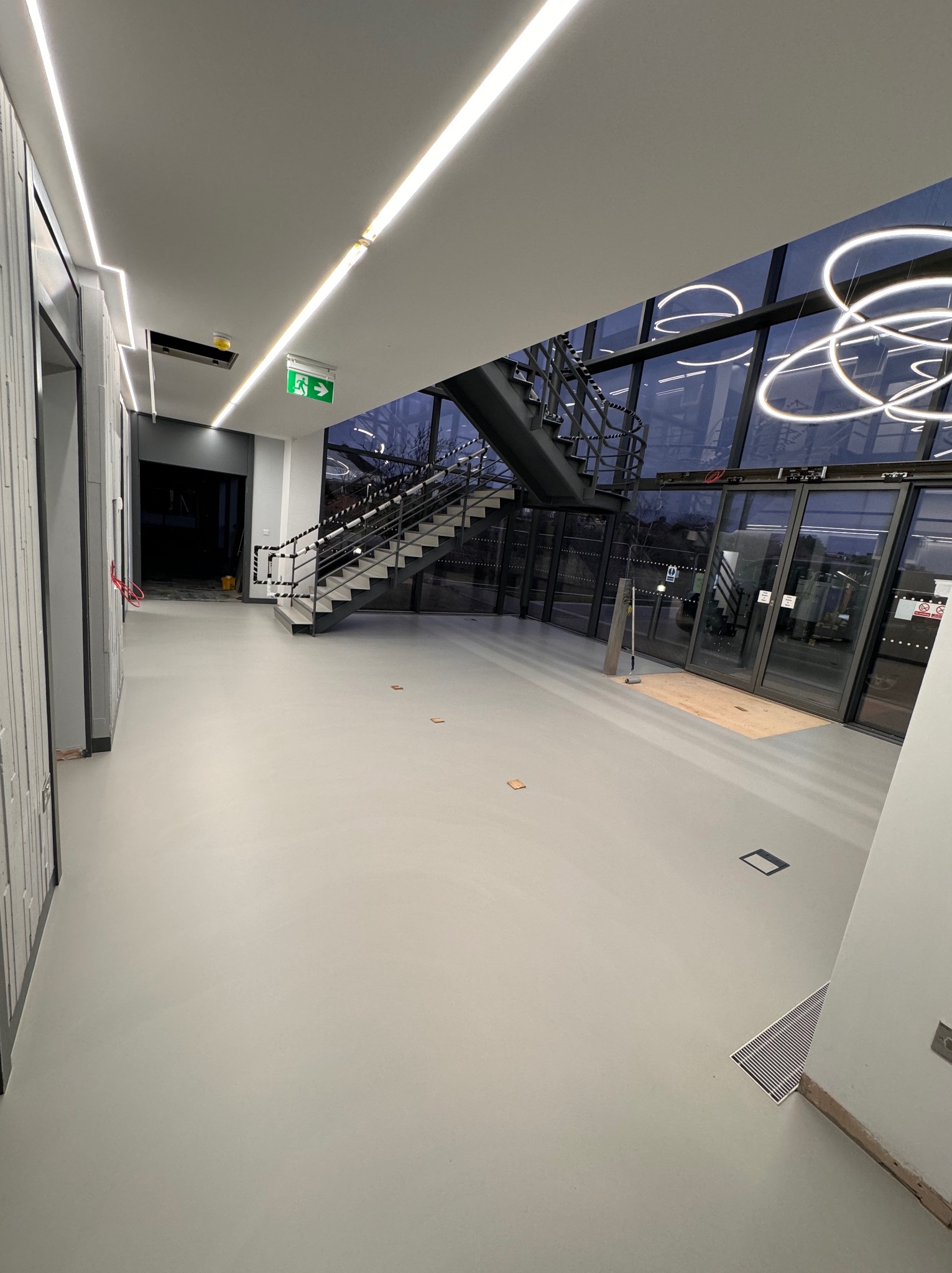 Wnętrze nowoczesnego budynku z szarą, gładką posadzką, widoczne schody, przeszklona ściana z dekoracyjnym oświetleniem i drzwi wejściowe.