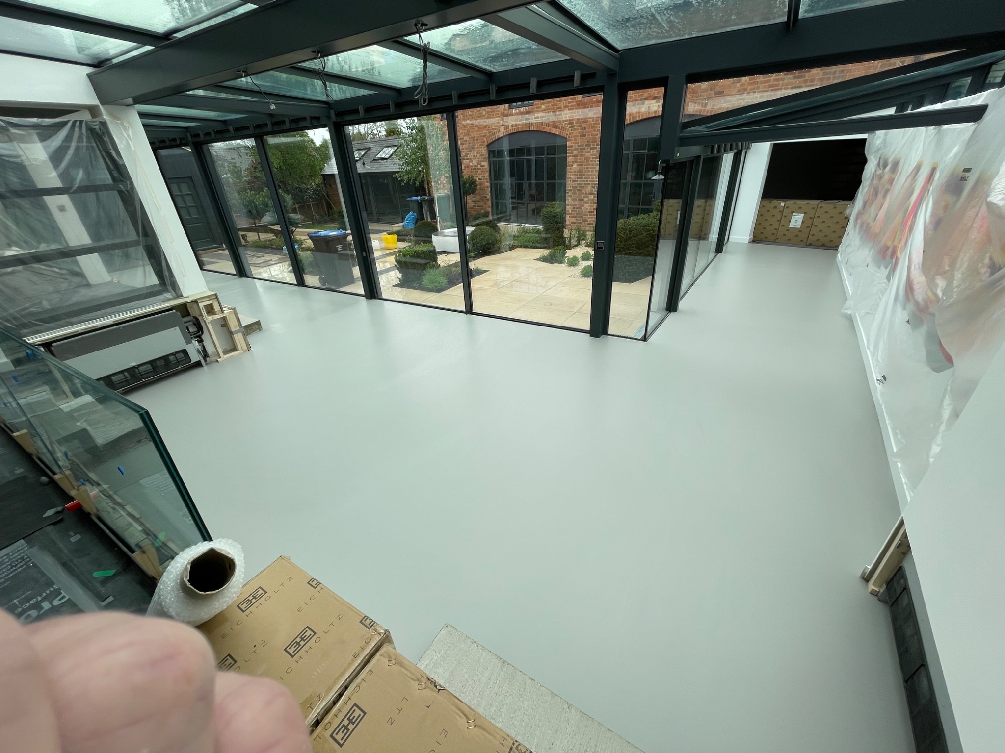 Jasnoszara, gładka posadzka w nowoczesnym, przeszklonym pomieszczeniu z widokiem na zadbany ogród. Elementy wykończeniowe zabezpieczone folią, widoczne kartony i materiały budowlane. Metalowa...