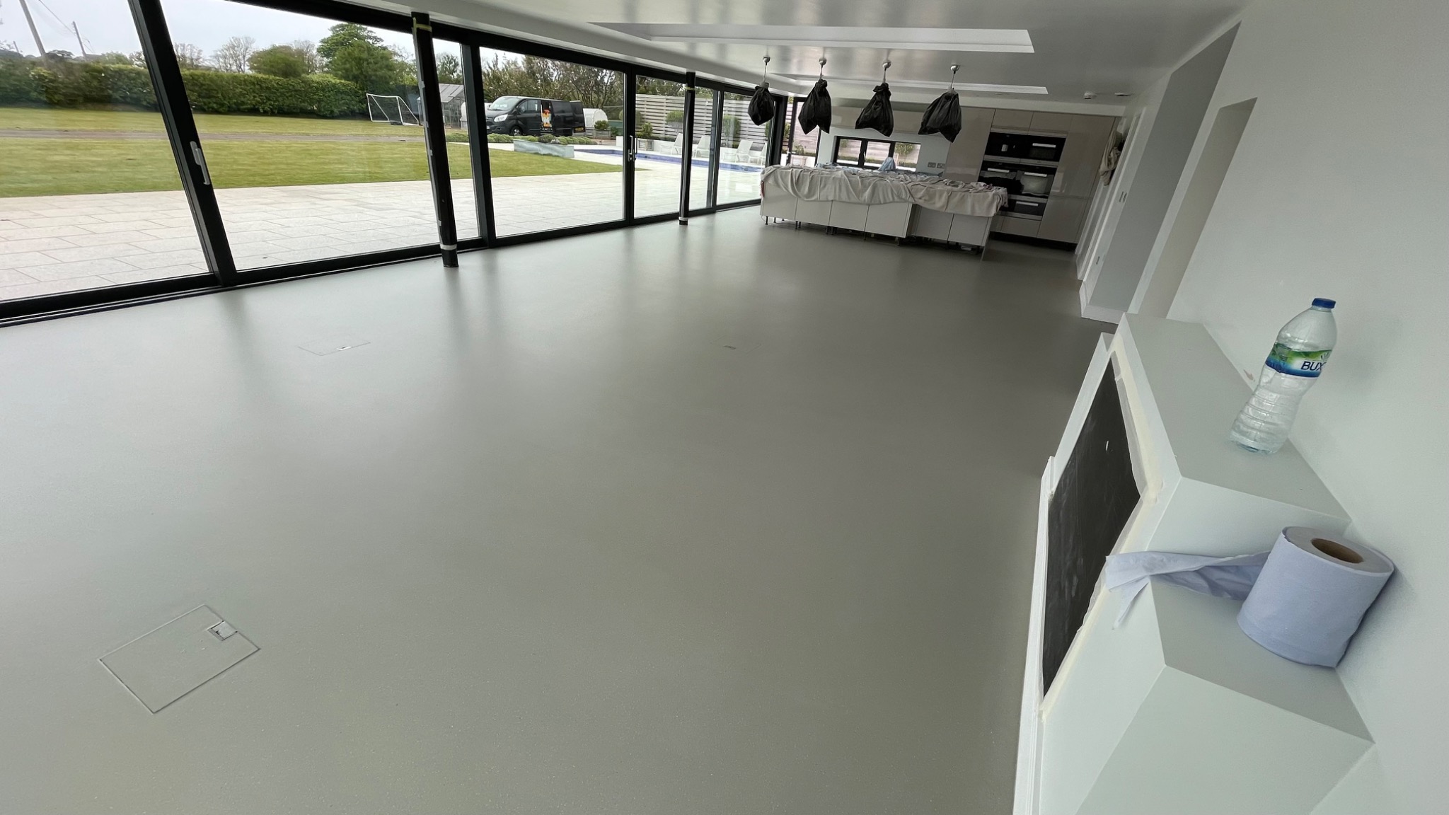 Jasnoszara, jednolita posadzka w przestronnym wnętrzu z dużymi przesuwnymi oknami wychodzącymi na zielony trawnik, widoczne klapy rewizyjne w podłodze, butelka wody i rolka papieru na białym...