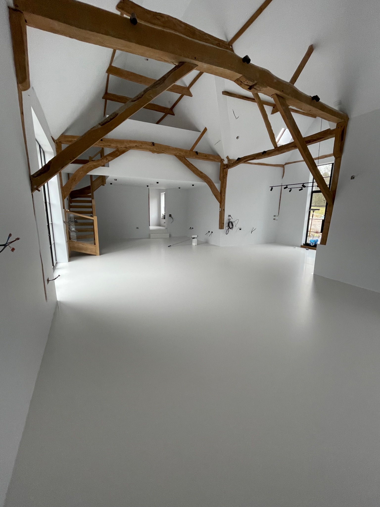 Jasne, przestronne wnętrze z białą, jednolitą podłogą, drewnianą konstrukcją dachu i schodami w tle, widać przygotowane puszki z farbą przy ścianie.