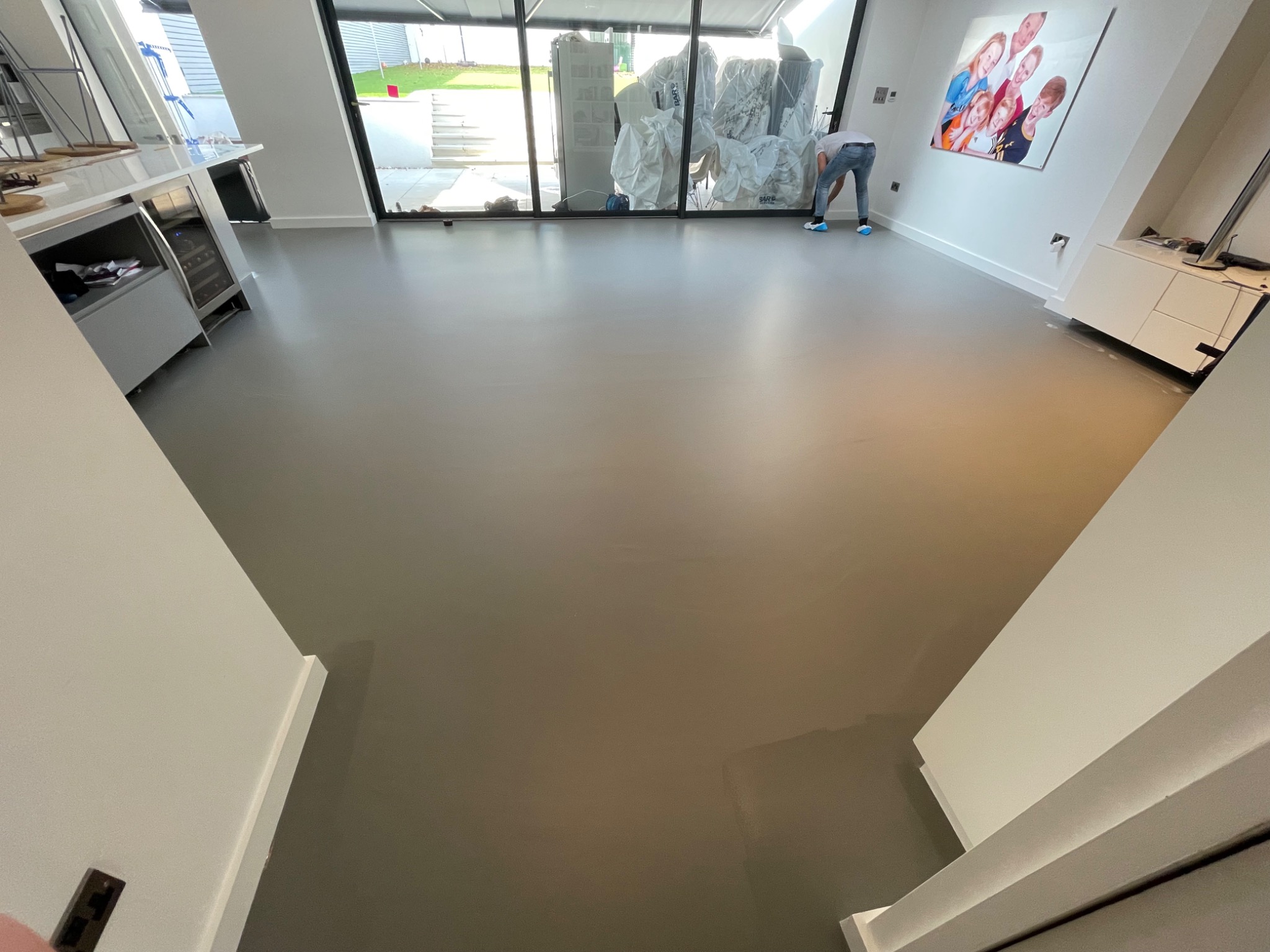Gładka, szara posadzka w nowoczesnym wnętrzu z dużym oknem, przez które widać zapakowane meble i pracownika w niebieskich ochraniaczach na butach, pochylonego nad podłogą.