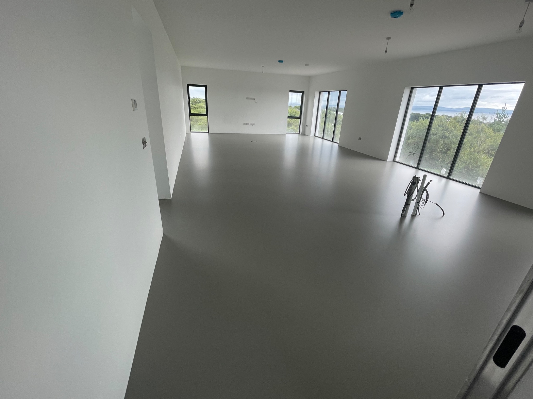 Szara, gładka posadzka w przestronnym, jasnym pomieszczeniu z dużymi oknami i widokiem na zieleń. Na podłodze widoczne narzędzia i drabina oparta o ścianę.