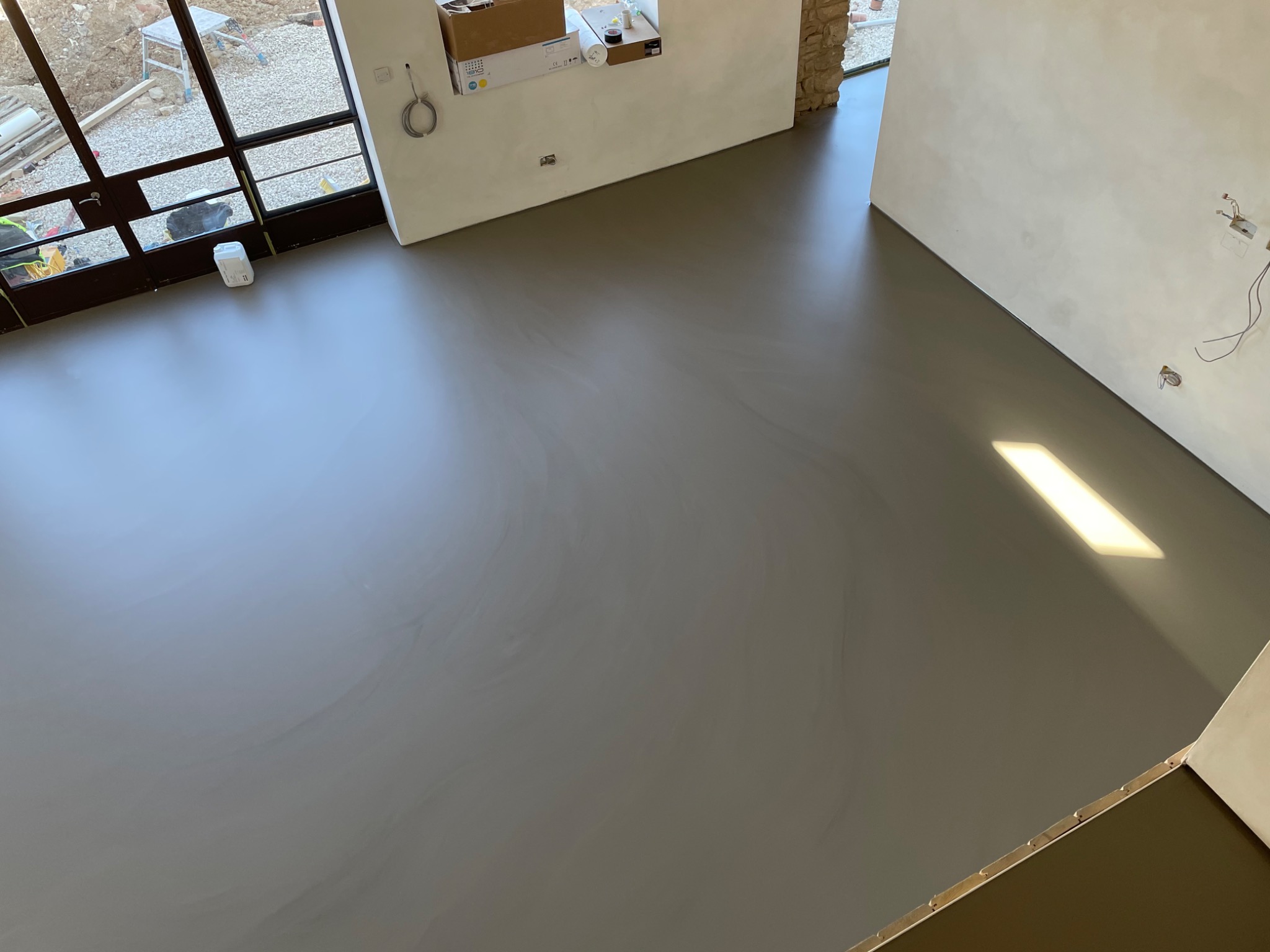 Świeżo wylana, gładka posadzka w szarym odcieniu, widok z góry, z oknem i drzwiami balkonowymi w tle, widoczne instalacje elektryczne w ścianie.