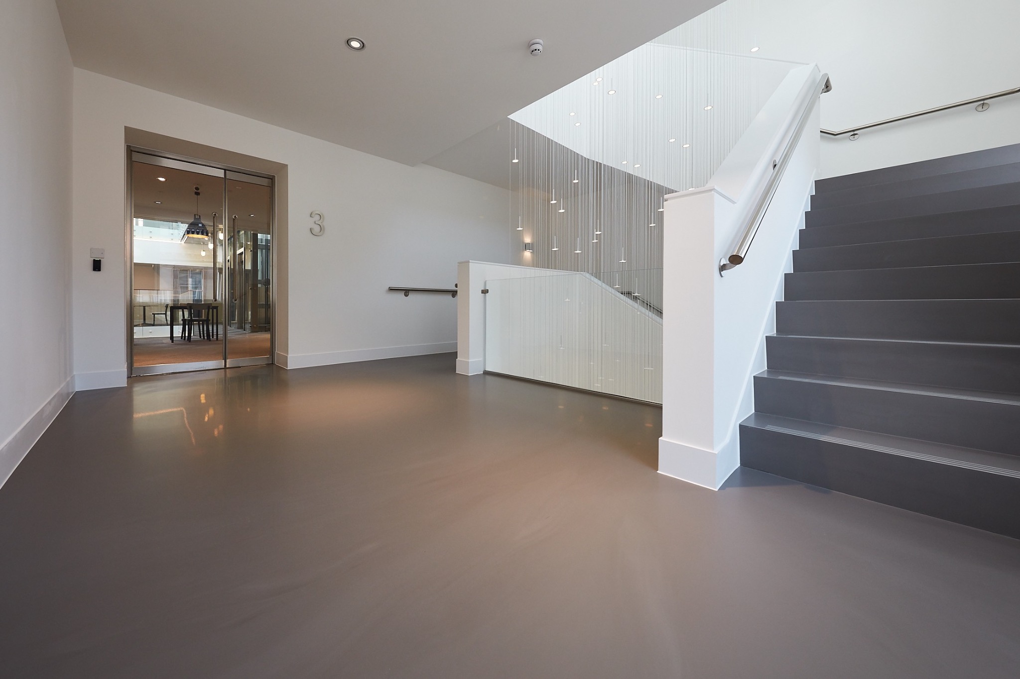 Szare, jednolite wykończenie podłogi w nowoczesnym holu z widocznymi schodami i przeszklonym wejściem, oświetlonymi wiszącymi lampami.