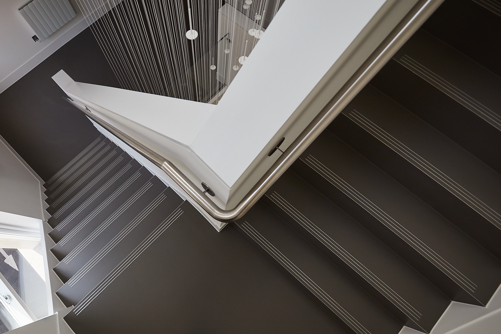 Nowoczesne schody z ciemną posadzką i metalową barierką, widok z góry na dół, z dekoracyjnym oświetleniem sufitowym.