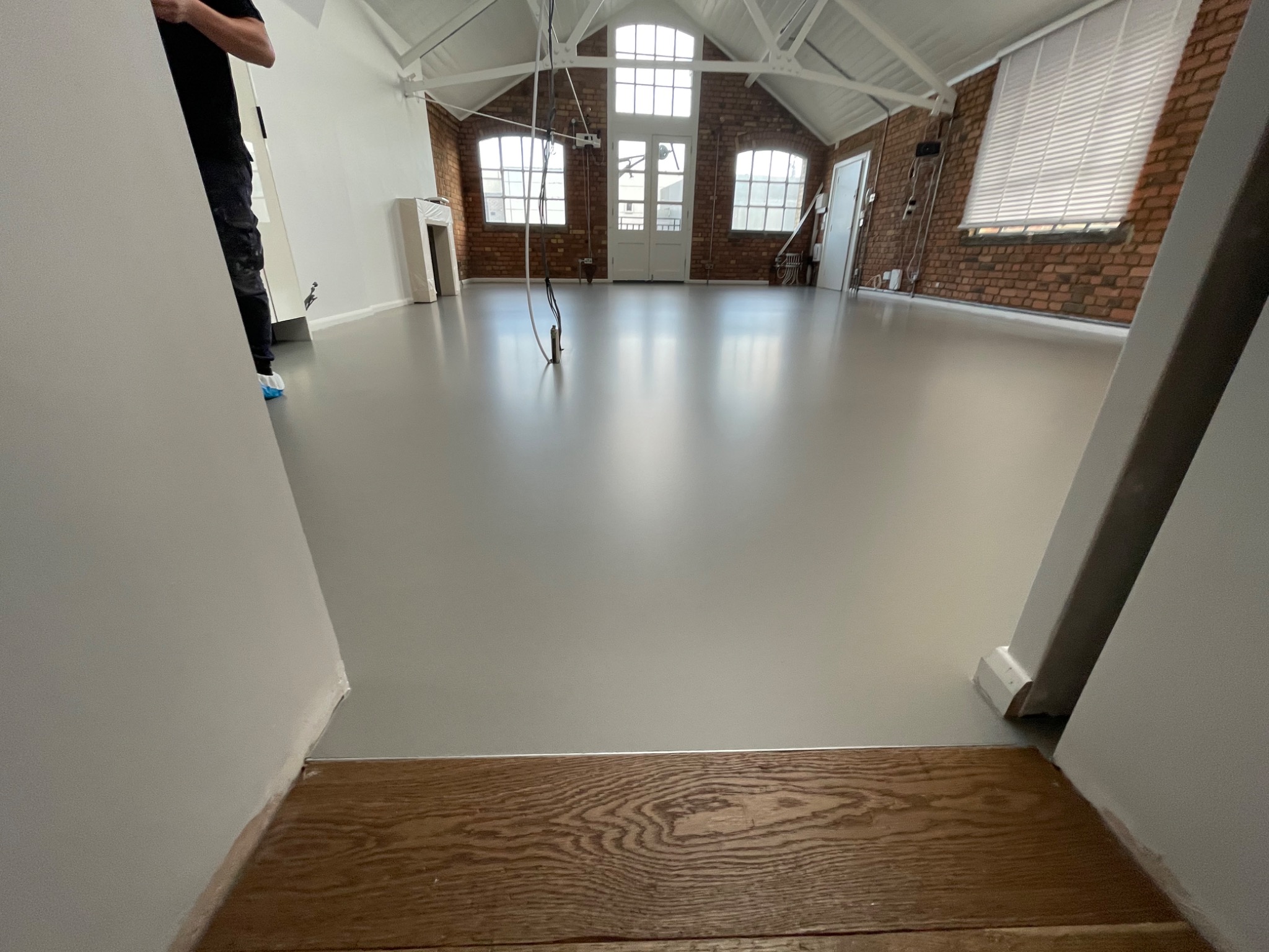 Jasnoszara, gładka posadzka w przestronnym, loftowym wnętrzu z ceglanymi ścianami i dużymi oknami. Widoczne elementy instalacji pod sufitem.