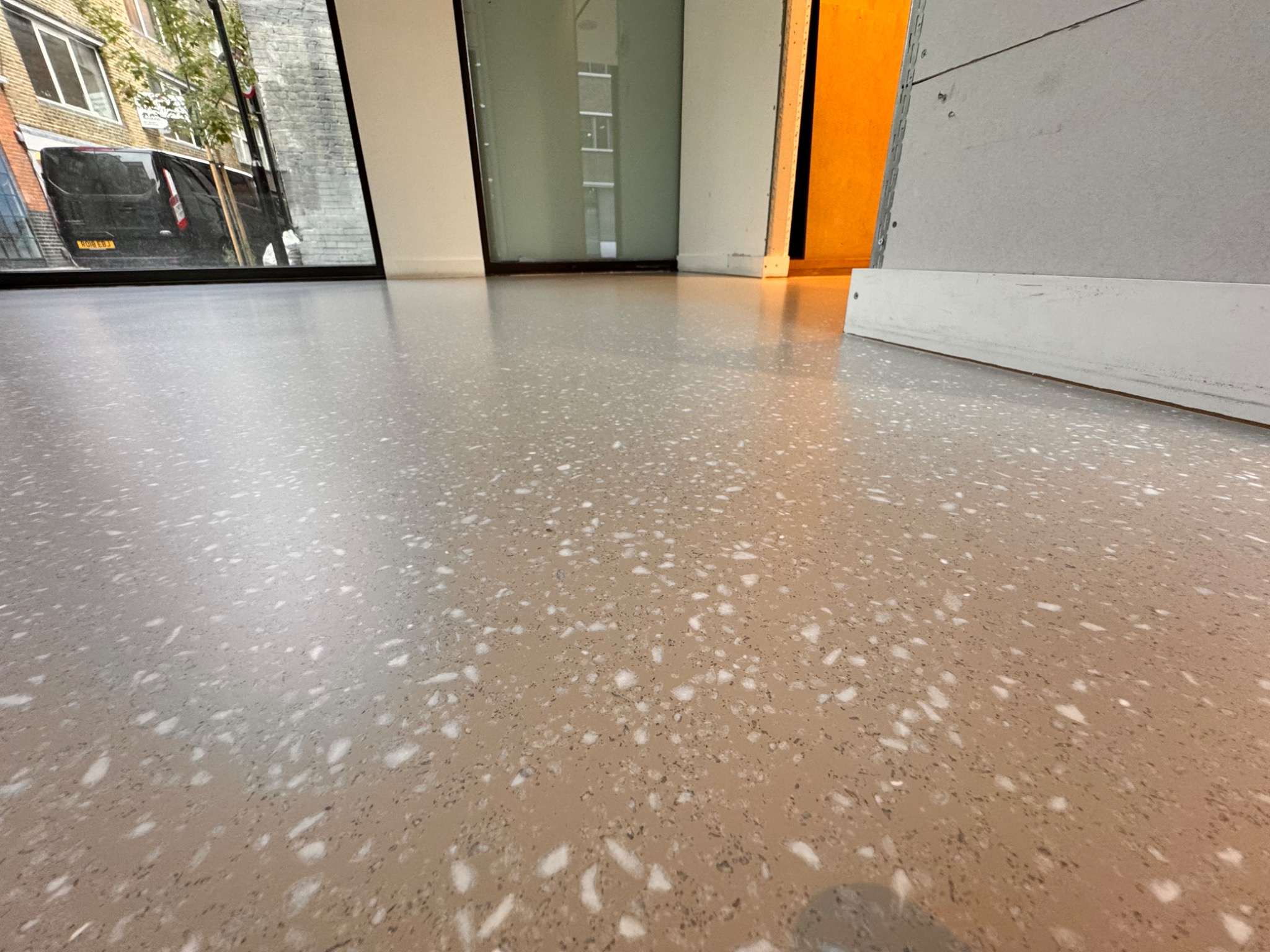 Beżowa posadzka żywiczna z białymi i brązowymi wtrąceniami, imitująca terrazzo. Widok z niskiej perspektywy, z odbiciem ulicy w oknie. Gładka, bezspoinowa powierzchnia.