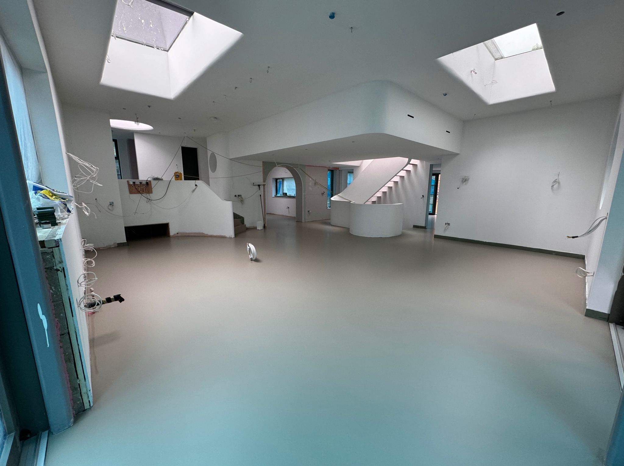 Jasnoszara, gładka posadzka w przestronnym, jasnym wnętrzu z oknami dachowymi. Widoczne schody, instalacje elektryczne i przygotowania do wykończenia.
