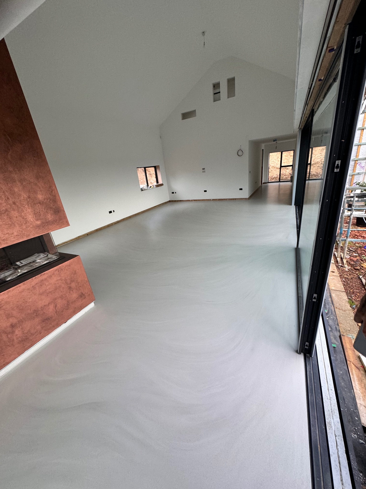 Jasnoszara, gładka posadzka w przestronnym salonie z kominkiem o miedzianym wykończeniu. Widoczne duże okna przesuwne i wysoki sufit ze skosami.