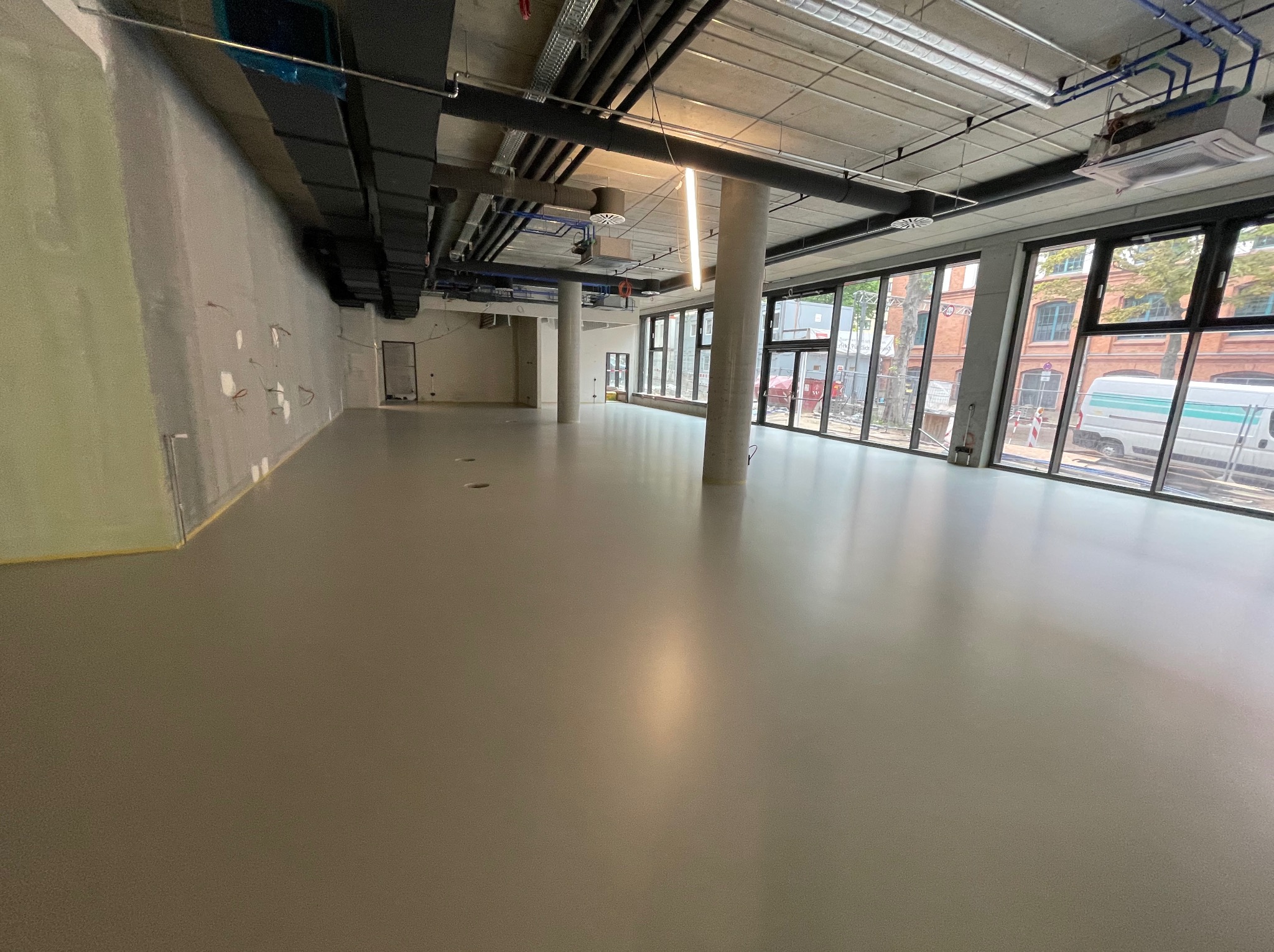 Jasnoszara, gładka posadzka w przestronnym, surowym wnętrzu z betonowym sufitem i widocznymi instalacjami. Duże okna wpuszczają światło dzienne.