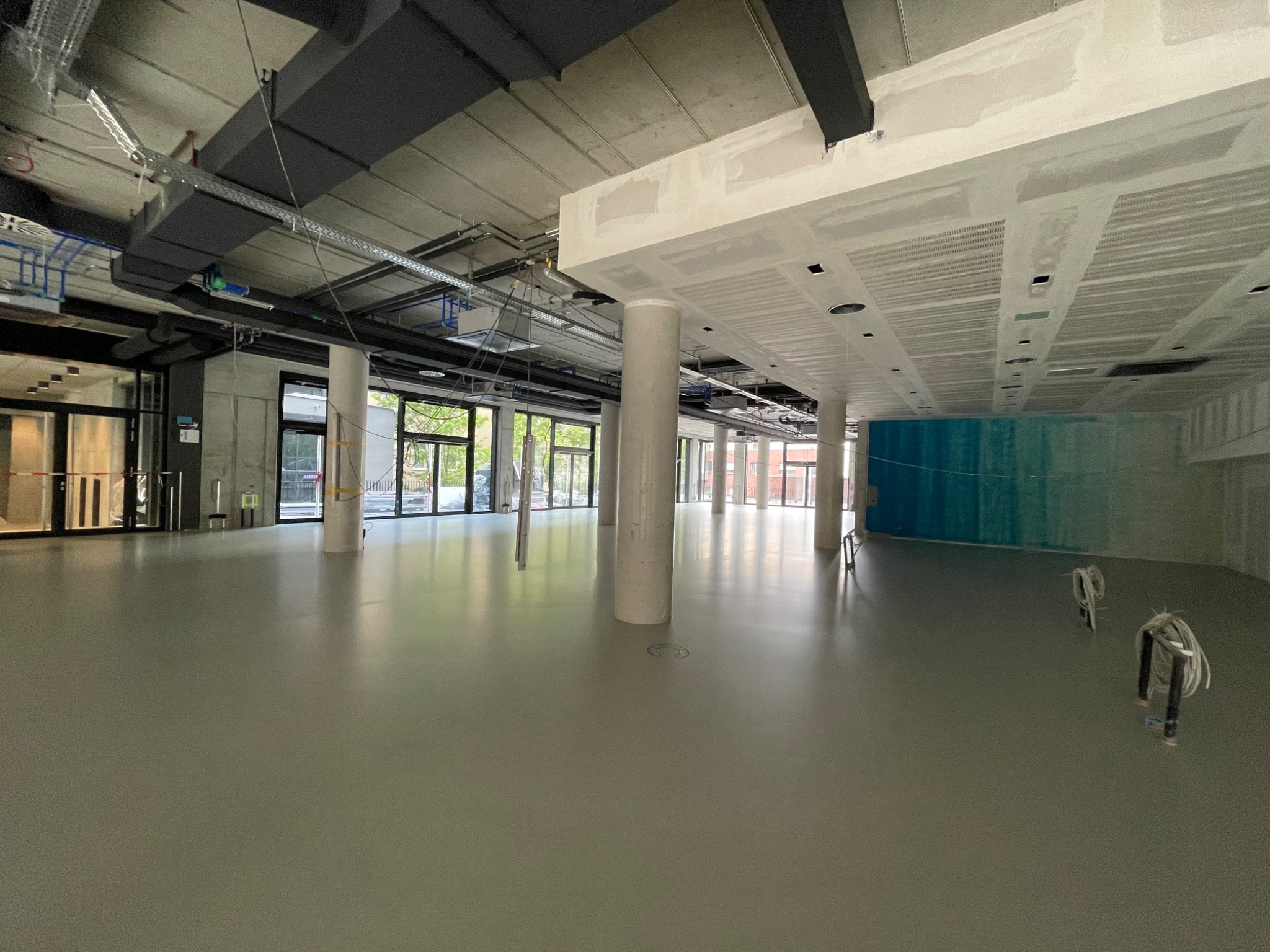 Jasnoszara, gładka posadzka w przestronnym, surowym wnętrzu z kolumnami i instalacjami pod sufitem. Widoczne kable i elementy wykończeniowe.