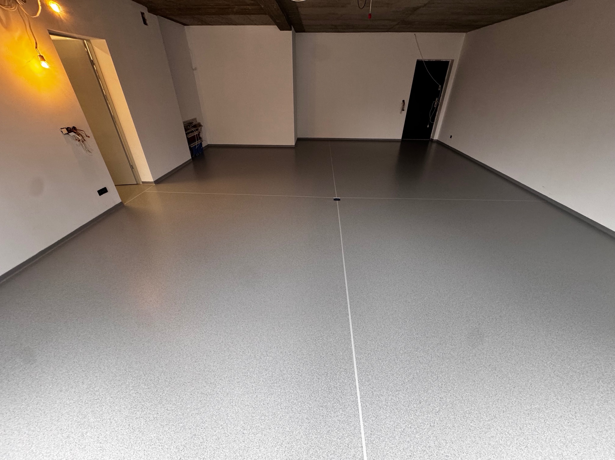 Szara, gładka posadzka żywiczna w surowym wnętrzu z betonowym sufitem. Widoczne białe ściany i czarne drzwi. Minimalistyczny design, surowy stan wykończenia.