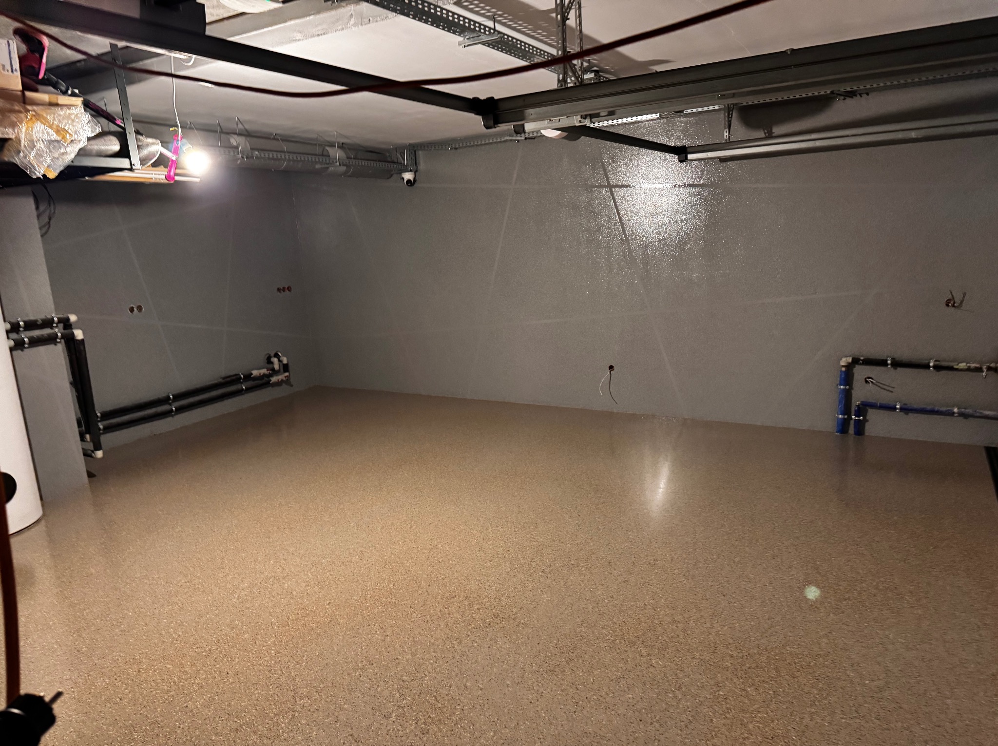 Jasna, gładka podłoga w garażu lub pomieszczeniu gospodarczym, z widocznymi instalacjami przy ścianach. Ściany szare, gładkie, z widocznymi elementami instalacji.