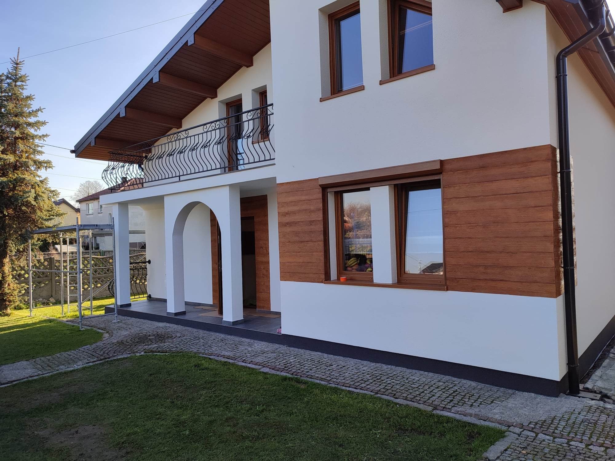 Biały dom jednorodzinny z balkonem i drewnianymi akcentami elewacyjnymi, widok na fragment elewacji i otoczenie z kostką brukową i trawą.