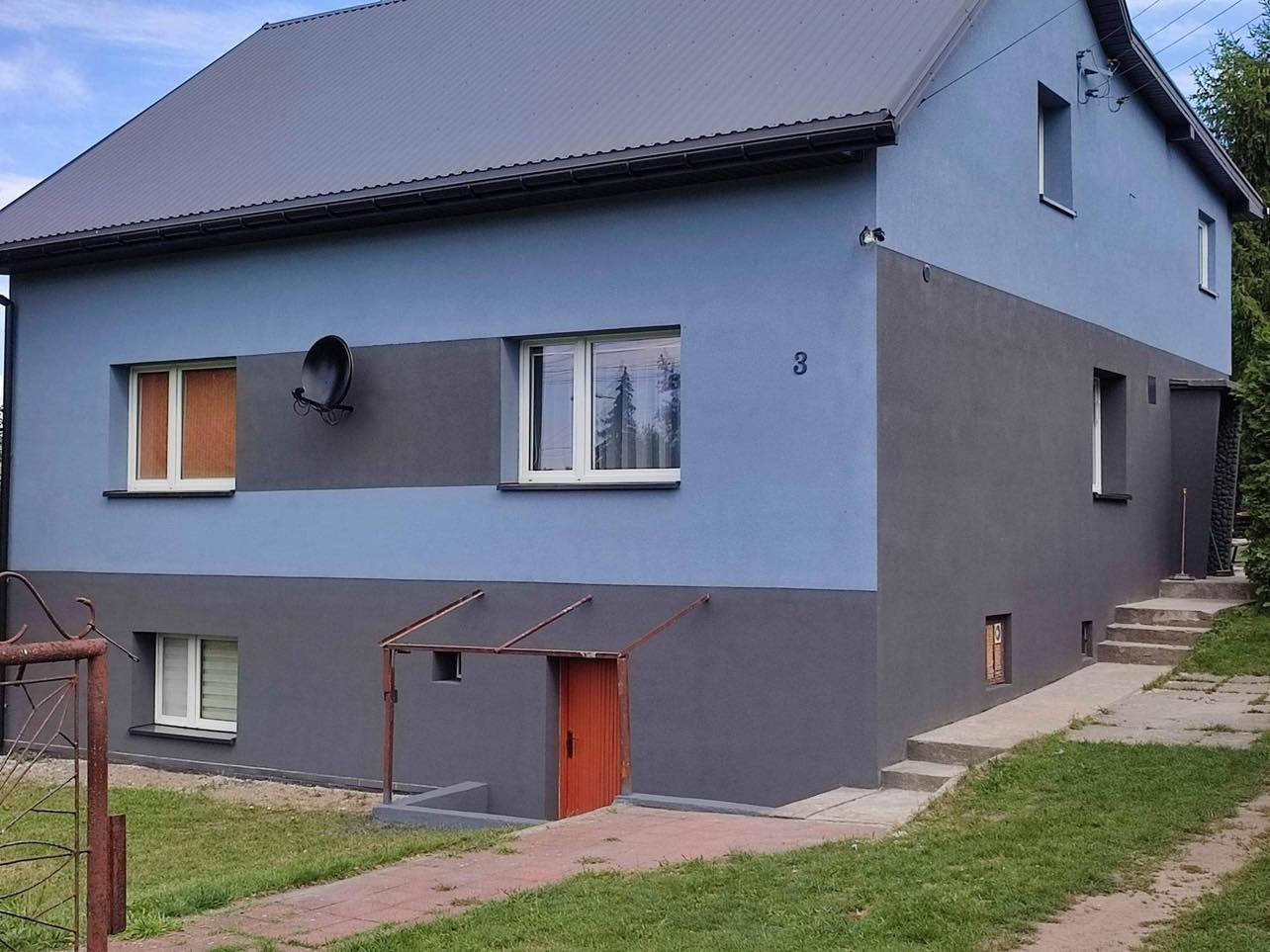 Dwukolorowa elewacja domu z szarą podmurówką i niebieską górną częścią, widoczna antena satelitarna i numer domu '3'.