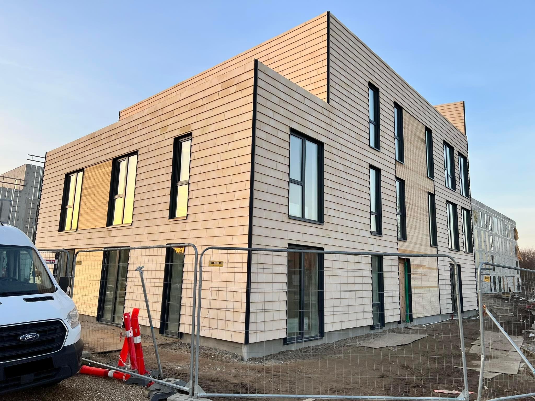 Nowoczesny budynek wielorodzinny z elewacją z drewnopodobnych paneli, czarnymi oknami i widocznym ogrodzeniem budowlanym oraz zaparkowanym białym vanem.