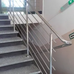 Polerowana, metalowa balustrada schodowa z poprzecznymi prętami, zamontowana na granitowych schodach w budynku z oznaczeniem drogi ewakuacyjnej na ścianie.