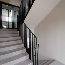 Klatka schodowa z szarymi, marmurkowymi stopniami i czarną, minimalistyczną balustradą, widok z dołu na górę, z widocznym fragmentem przeszklonego wejścia.