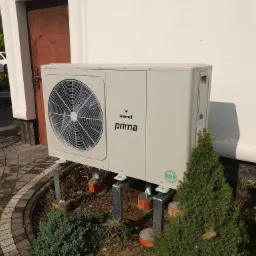 Zewnętrzna jednostka pompy ciepła Galmet Prima R32 zamontowana na metalowych wspornikach z betonowymi podstawami, obok ozdobnych krzewów i ścieżki z kostki brukowej.