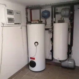 Dwa białe, cylindryczne zbiorniki wodne w kotłowni, z widocznymi rurami, zaworami, manometrami i naczyniami przeponowymi, zamontowane na tle białych ścian i szarej podłogi z płytek.