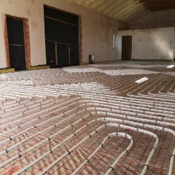 Rozłożone rury ogrzewania podłogowego na folii z siatką w surowym wnętrzu budynku w trakcie montażu, widoczne kolektory i pianka na suficie.