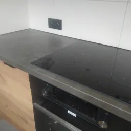 Fragment kuchni z drewnianą szafką, szarym blatem, czarną płytą indukcyjną Amica i piekarnikiem, widoczne białe płytki na ścianie i podwójne gniazdko elektryczne.